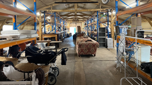 A wide shot of the inside of a barn which is now being used to house donated items. There is a row of sofas in the middle and to the left there are a row of prams. Either side are rows of high shelves with donated items on.