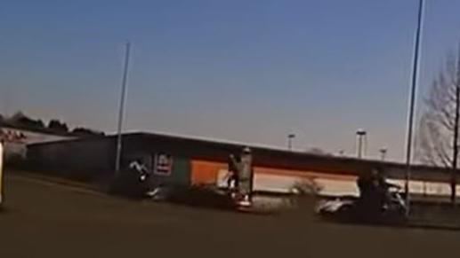 A still from dashcam footage showing a road junction on an industrial estate with a red convertible car stationary and a figure standing on the bonnet