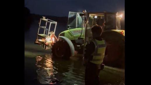 A police officer standing on the shore of Windermere while a tractor enters the water. It is dark outside.