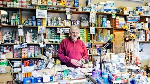 A man in a red top stood behind the counter in a shop selling electrical goods 