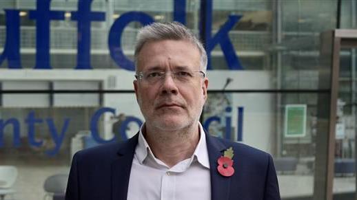 Paul West stands outside Suffolk County Council headquarters. He is not smiling. He has short grey hair and wears a navy suit with a white shirt underneath.