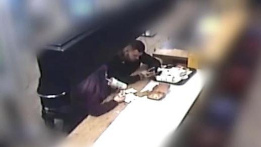 A screenshot taken from CCTV footage inside a McDonald's restaurant. The background is blurred so you can only see the pair sitting at a table. Behind them is a black pushchair with the baby inside. Reilly is drinking from a fast-food cup while Kember searches on his phone. A tray of food packaging is on the table in front of them.