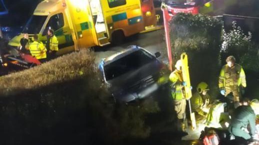 A silver VW Passat poking through a hedge it has crashed through. Behind it are ambulances. In the foreground firefighters and a paramedic