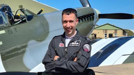 Sqn Ldr Mark Long, with short brown hair and wearing a black flight suit, smiles while folding his arms. Immediately behind him is the wing of a Spitfire.