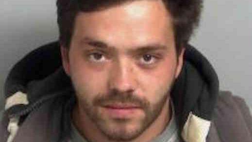 Charles Benson-Clark wearing a grey crew-neck shirt layered under a dark jacket, seated against a plain light grey background. He has black hair.