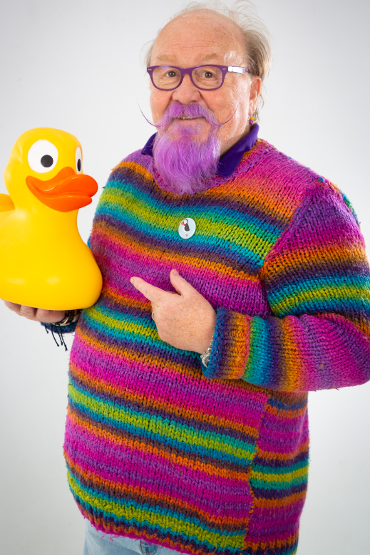 Man with glasses and purple beard. He is holding a large yellow rubber duck in one hand and is pointing at it with the other. He's wearing a brightly striped wool jumper.