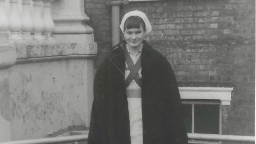 Image shows black and white photo of Dawn Wintle smiling into the camera, with a brick-building in the background, and wearing her uniform. She wears a black coat with her hair pulled back and a white nurse cap.