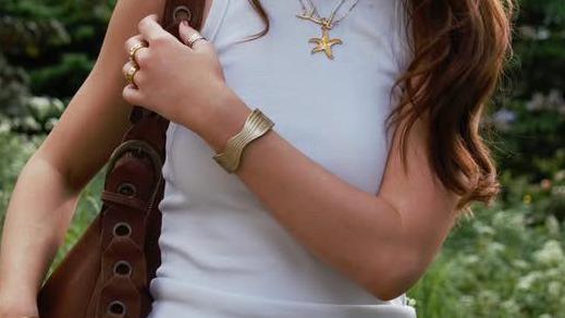 A promotional image for the bracelet. It shows a slim woman, between her shoulders and waist, holding her arm over her front while she pulls her handbag up onto her shoulder. The chunky gold bracelet is front and centre. She wears a white vest top, and behind her is a field.