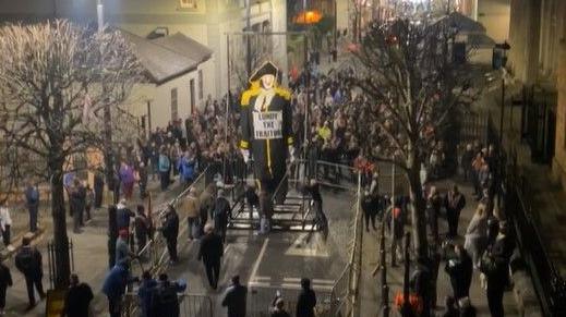 A large crowd around an effigy of Lundy. He is in a black uniform with white face and is wearing a sign reading: 'Lundy The Traitor'