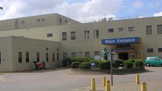 The main entrance of Milton Keynes General Hospital with some yellow bollards in the foreground and a roundabout with a statue in the middle of it directly outside the entrance.