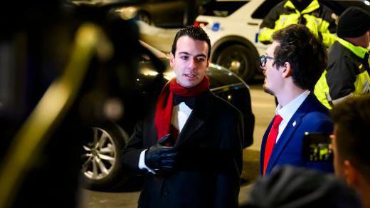 Paul Ingrassia stands outdoors with a coat on, talking to another man