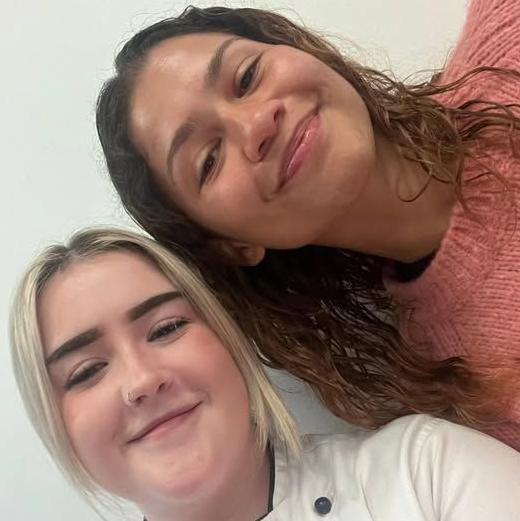 Zendya has brown wavy hair, a pink jumper and is smiling as she poses for a selfie with Joss. Joss has blonde hair and is wearing an ice cream shop outfit.