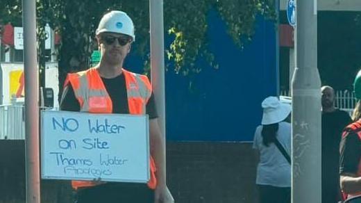 A water worker holding a whiteboard that says "No water on site. Thames Water apologises". He is wearing a hi-vis vest and a white helmet.