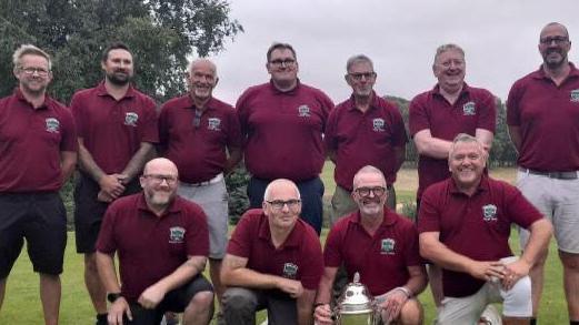 The Beccles Golf Club team smile after winning their competition. There are 11 men all wearing the same red polo T-shirt. Stuart Hagger sits on the bottom row crouching. He hold the trophy.