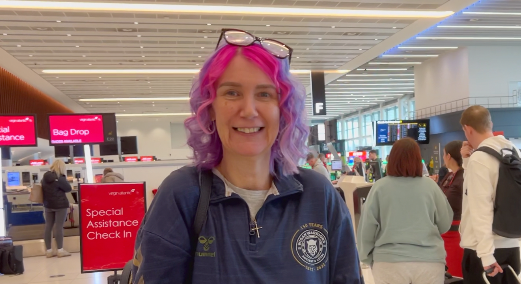 Lindsay smiles at the camera, she wears a blue jumper with a grey t-shirt underneath. Se has pink and purple shoulder-length wavy hair and glasses on her head.