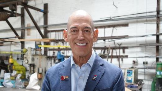 Roly Woods is dressed smartly in a navy jacket, with poppy badges, and a blue checked shirt. He is grinning at the camera and standing in the forge.