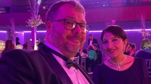 A close-up of a man with a beard, wearing glasses and a suit with a bow tie. He is pictured alongside a smiling woman wearing a necklace and dress