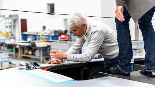 Geoff Willis with short white hair, wearing a grey sweater, leans on part of a grey hull in a test room. A pair of legs in blue trousers is visible to his left.