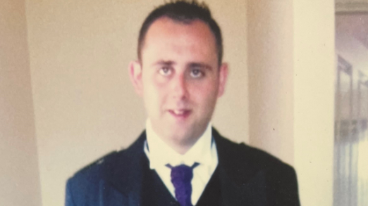 Head and shoulders shot of a pale looking man with short dark hair. He is smiling and looking at the camera. He is wearing a dark kilt jacket, a white shirt and a purple neck tie. The man appears to be standing in a domestic setting against a white wall.