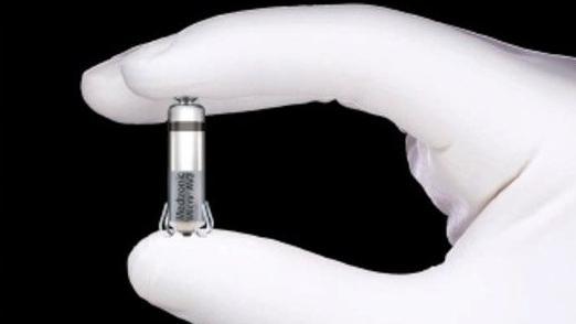 A gloved hand is holding the pacemaker between their thumb and index finger. It is very small and has a silver casing. 