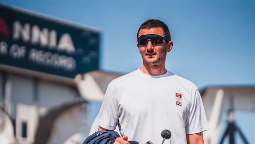 Giles Scott with short dark hair wearing a white T-shirt with a small union jack logo, on the Ineos Britannia