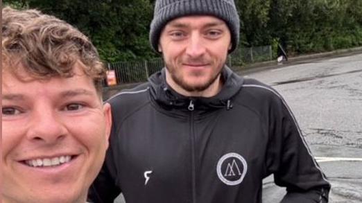 Will Fraser-Coombe is wearing a grey beanie hat, has short brown stubble and is wearing a gym-branded long-sleeved training jumper. Standing next to him is Grant Grego, who has light brown long hair and is wearing a grey hoodie. 