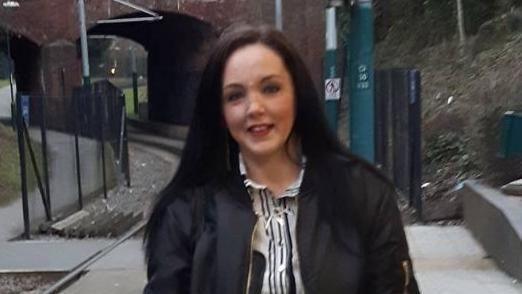 Ellen Marshall with dark brown long hair and wearing a black bomber jacket over a monochrome striped dress. She is standing on a train platform with the train tracks visible on the left