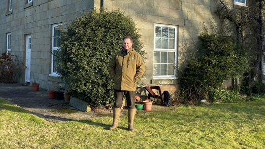 Louis Fell is standing outside a house. He's a man in his 40s wearing a green jacket and wellington boots. 