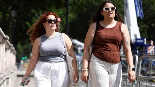 Two women walking through a street, with metal barriers to the right of them, wearing white trousers and vest tops - they both have sunglasses on.