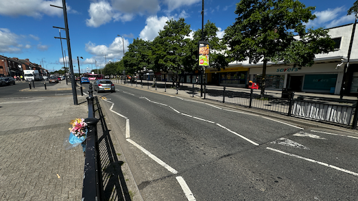 A road through a town centre with shops on one side. On the other side there is a bunch of flowers on the railings