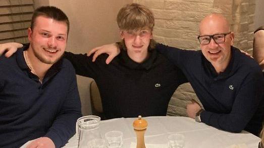 Harry, Freddie and Alain sat with their arms round each other at a table in a restaurant, with glasses on the table and a salt or pepper shaker.