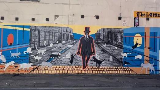 A view of a mural on a building. It shows a man in a black coat and hat surrounded by cats.