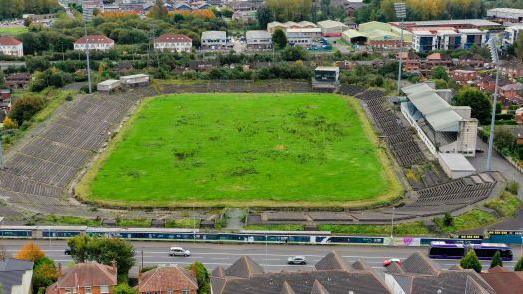 A bird's eye view of Casement Park stadium in Belfast showing an overgrown pitch in the centre of the frame, dilapidated seating around the park and rundown stands to the right of frame. In the background, residential housing.