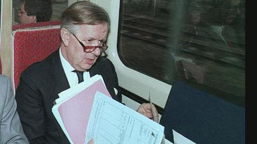 A man with papers in his hand wearing a suit and glasses is sitting on a train which has an interior from the 1970s.