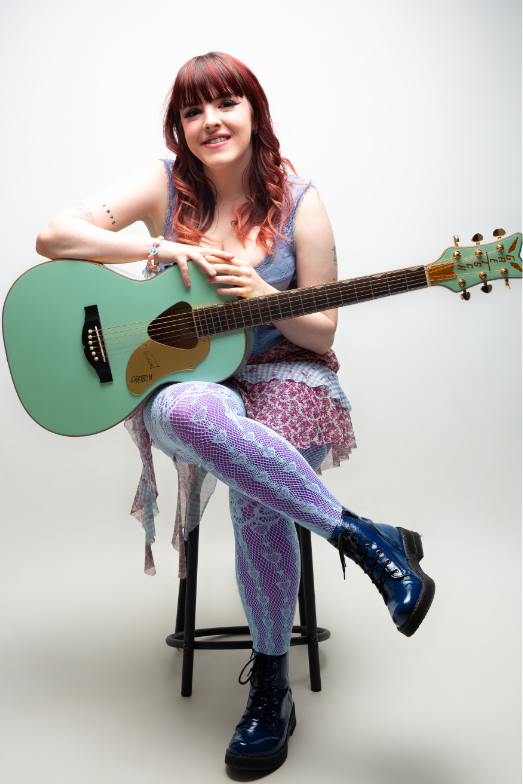 Young woman with long red hair is sitting on a stool with a green guitar. She's wearing lacy blue and purple tights. 
