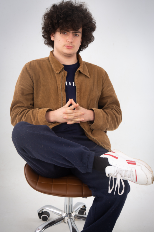 Boy with curly black or dark brown hair. He's sitting on a stool with one leg crossed. He's wearing a brown jacket, black t-shirt, navy trousers and white trainers. He's crossing his hands in his lap.