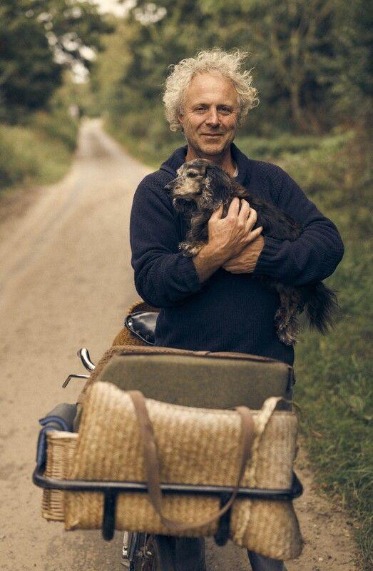 Charlie Mackesy is standing by his bicycle which has luggage at the front of it. He wears a blue jumper and holds a small dog.