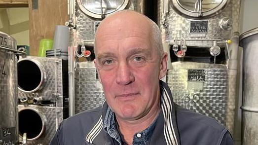 A picture of Robb Capp. He is facing the camera and smiling. He is wearing a blue overall on top of a blue shirt. He is bald. He is standing in front of large vats and other machinery at the vineyard.