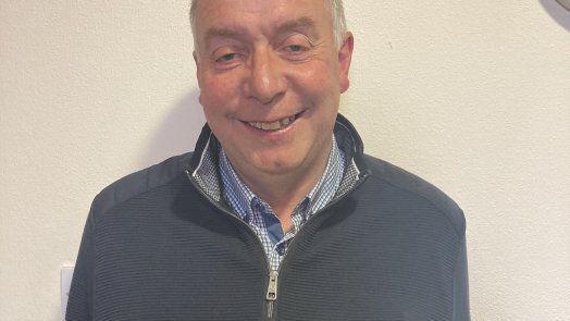 Mr Lynch has short grey hair and is smiling at the camera as he wears a navy half-zip and checked blue shirt. He is standing in front of a cream wall.