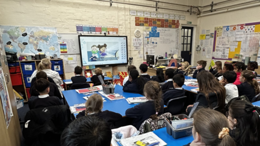 A school classroom with children sitting at desks looking at a television mounted on the wall