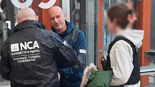 Arsen Feci being extradited by two NCA officers. One officer is wearing a black windbreaker while the other is in a grey hoodie and black vest over the top. Feci is wearing a blue hooded top.