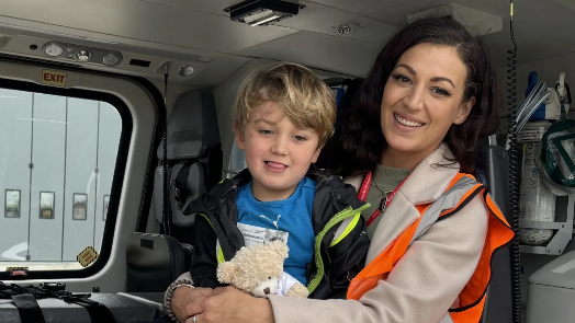 A young boy with fair hair sits with a woman with dark hair wearing a hi-vis jacket in a stationary helicopter.