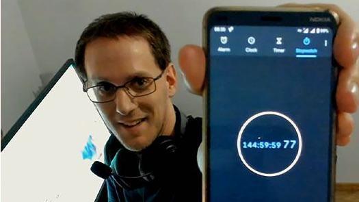 A man wearing glasses, holding a phone to the screen which reads "144:59:59:77".