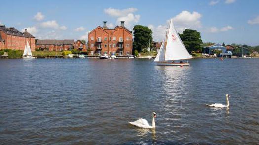 The calm water of Oulton Broad with two swans in the foreground and sailing boats and large red brick buildings in the background