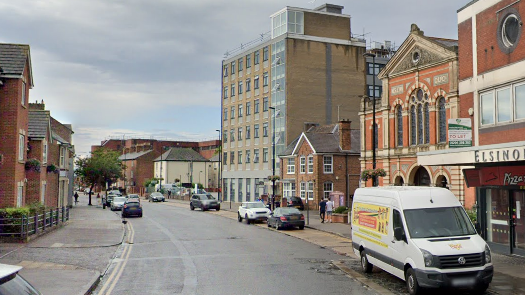 A general view of Buckingham Street in Aylesbury. Vehicles are parked either side of the road while buildings including flats and a parade of shops line either side of the road.