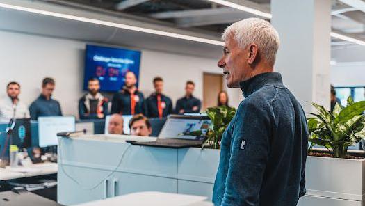 Geoff Willis wearing a blue top talks to a line of people, some wearing red ties, in an office with a laptop on a shelf nearby alongside pot plants.