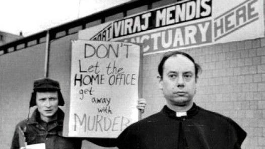 Father John Methuen is standing in front of the Church of the Ascension in Manchester. On the building are the words: "Viraj Mendis sanctuary here."
Behind Father Methuen is a man holding a sign which reads "Don't Let the Home Office get away with MURDER".