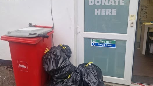 A pile of black binbags leaning against a red plastic wheelie bin, outside a white UPVC door with a "Donate Here" and a fire exit sign.