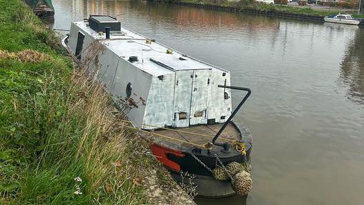 A narrowboat with rough white paint covering the top of the boat while the hull of the vessel remains red and black. The boat sits on muddy brown water in a canal with the grassy side of a path next to it.