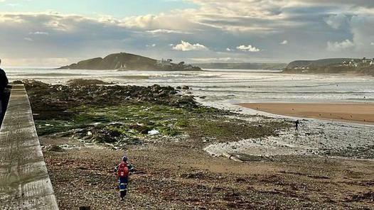 A beach with rocks on the left covered with seaweed and an island in the distance with an estuary flowing onto the beach.